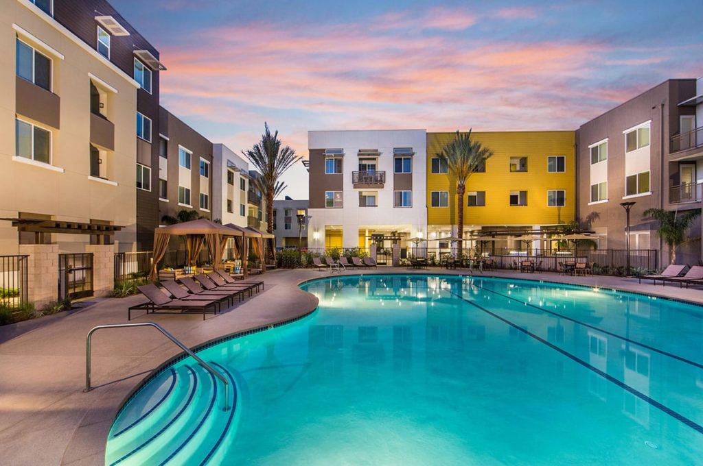 Lit blue pool with lounge chairs on either side and cabanas in the background surrounded by white, yellow and brown apartment buildings
