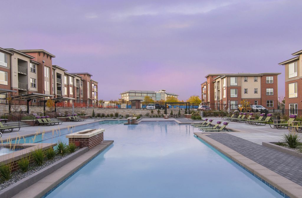 Blue long pool with green lounge chairs on the deck and red and brown apartment buildings surrounding on a sunset sky