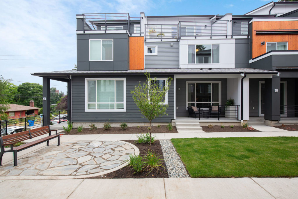 Three story gray townhouse with orange wood paneling and a rooftop patio, with landscaping and a bench in front
