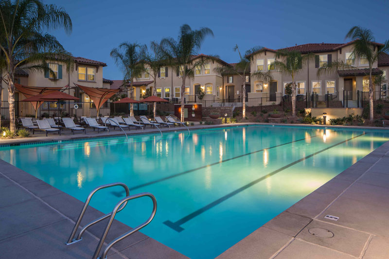 Lit-up pool at nighttime with white lounge chairs under red cabanas with cream-colored apartment buildings in the background