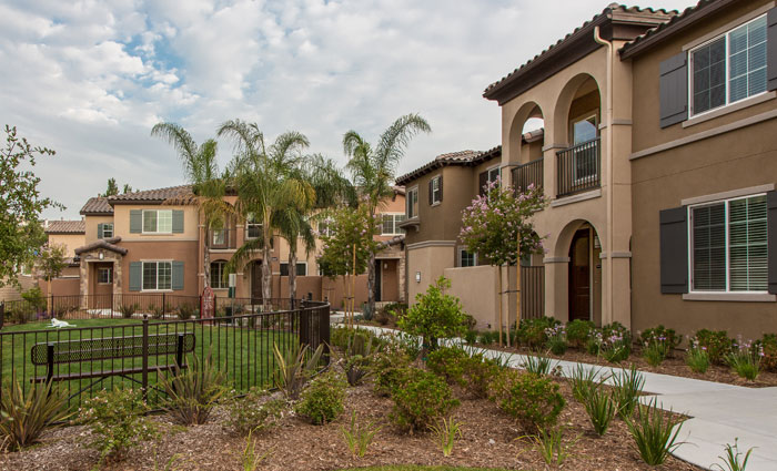 Light and dark brown two story townhouses surrounding a closed-in green space with a metal bench and mulch beds around