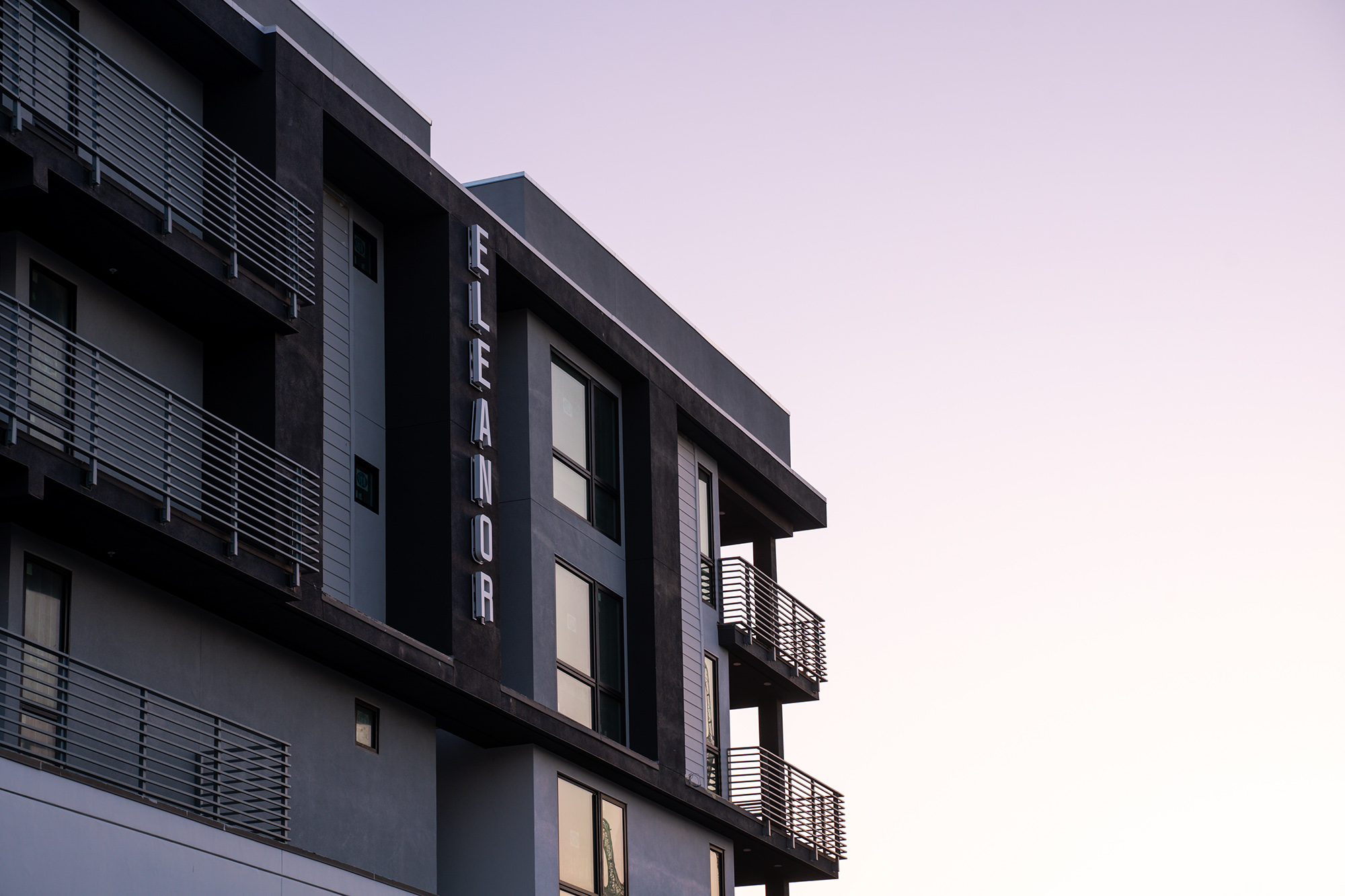 Corner of top of black and dark gray apartment complex with Eleanor letters on the side and balconies on the corner on a sunset sky