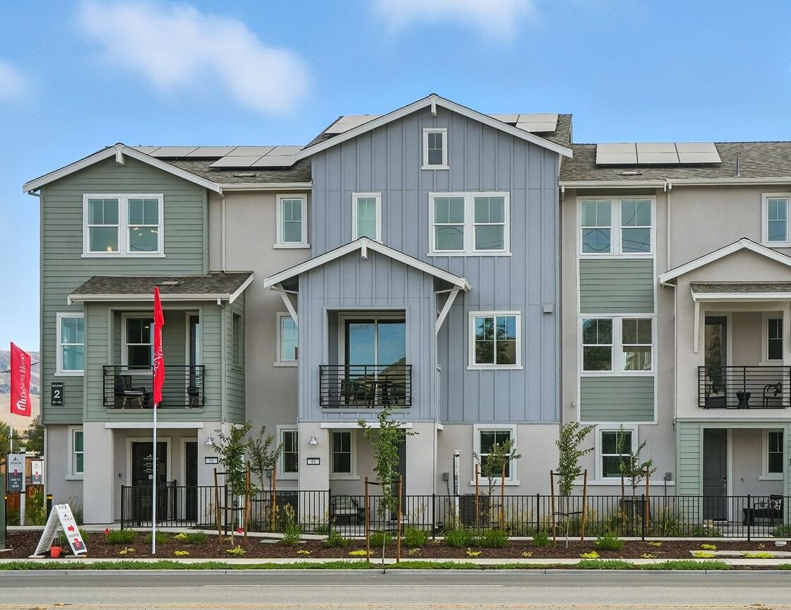 Three-story modern townhouse building with balconies and solar panels, facing a main road with mountains visible in the background under a blue sky.