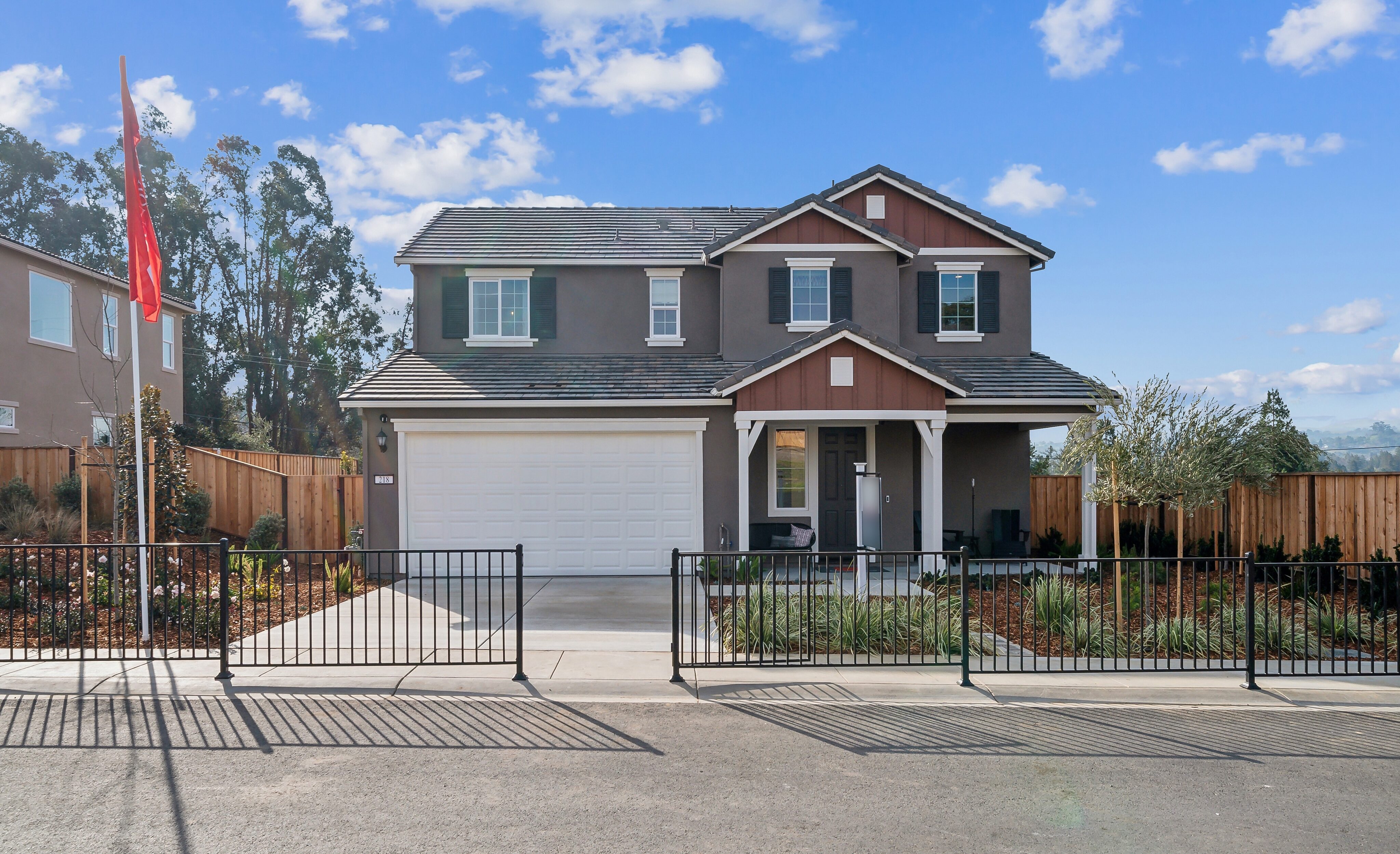 Two-story suburban house with a gray exterior, white garage door, and small front porch, surrounded by a black metal fence and landscaped yard under a blue sky.