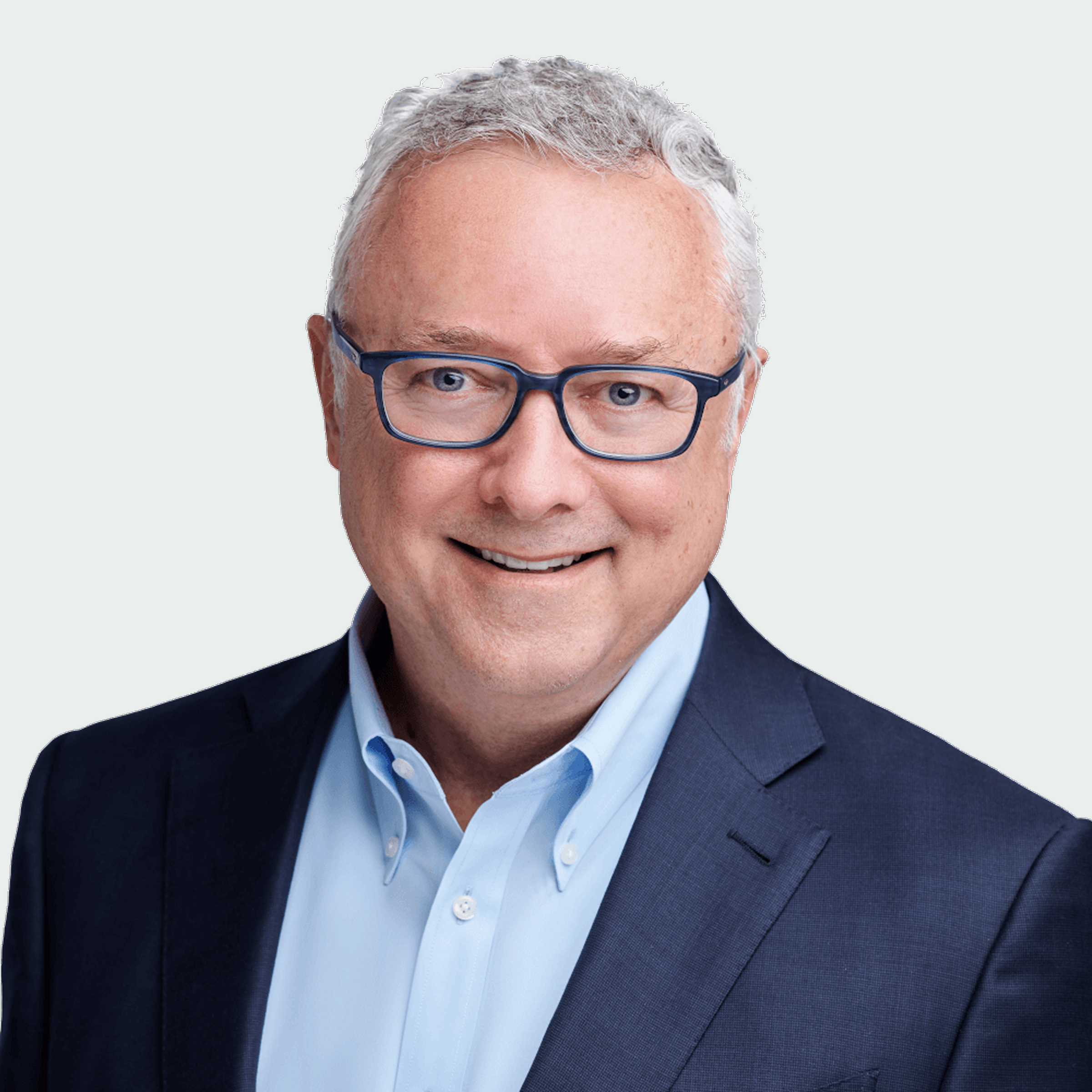 A man with gray hair and glasses, wearing a dark suit jacket and light blue dress shirt, smiles at the camera against a plain light background.
