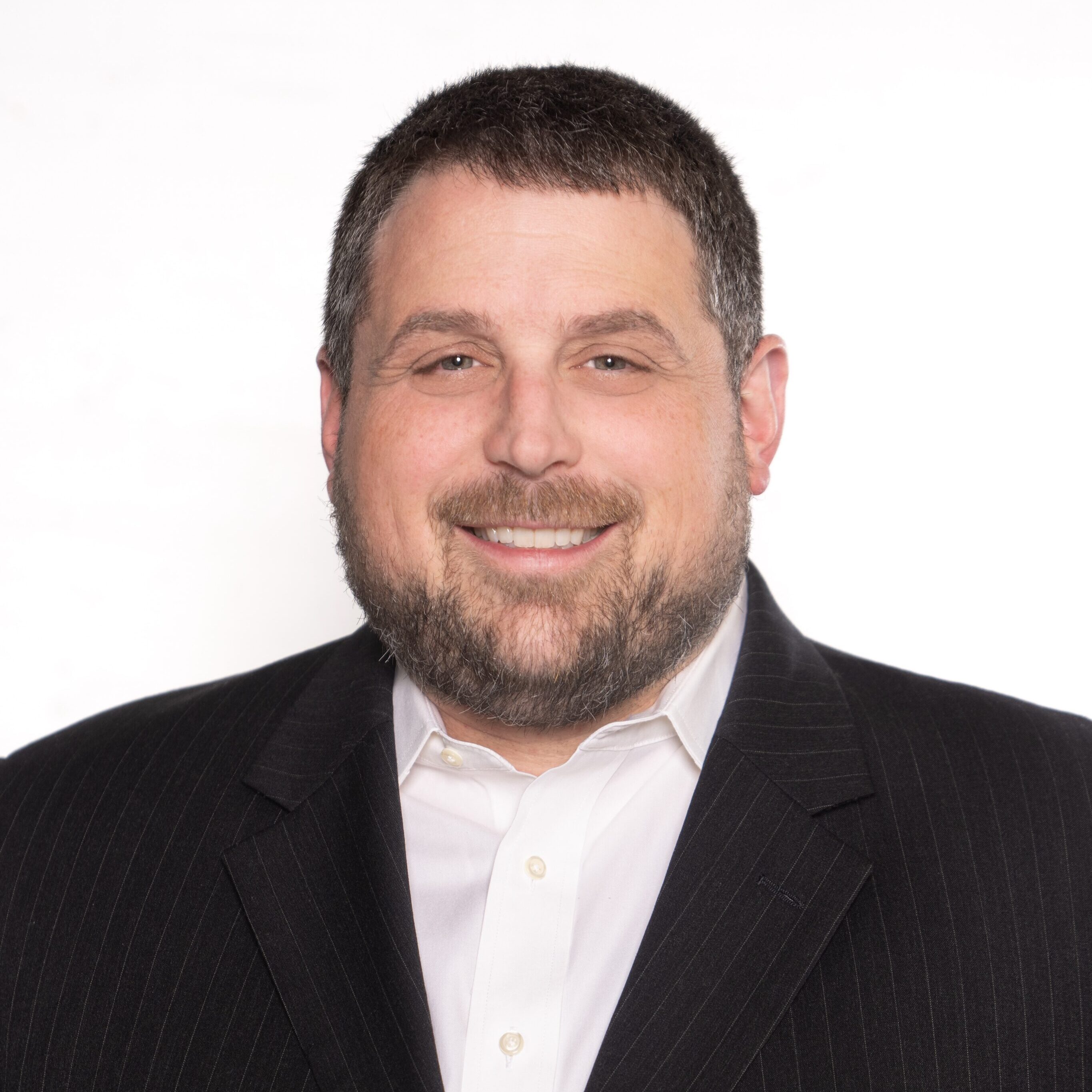 A man with short hair and a beard is wearing a dark pinstripe suit jacket over a white button-up shirt, smiling at the camera.