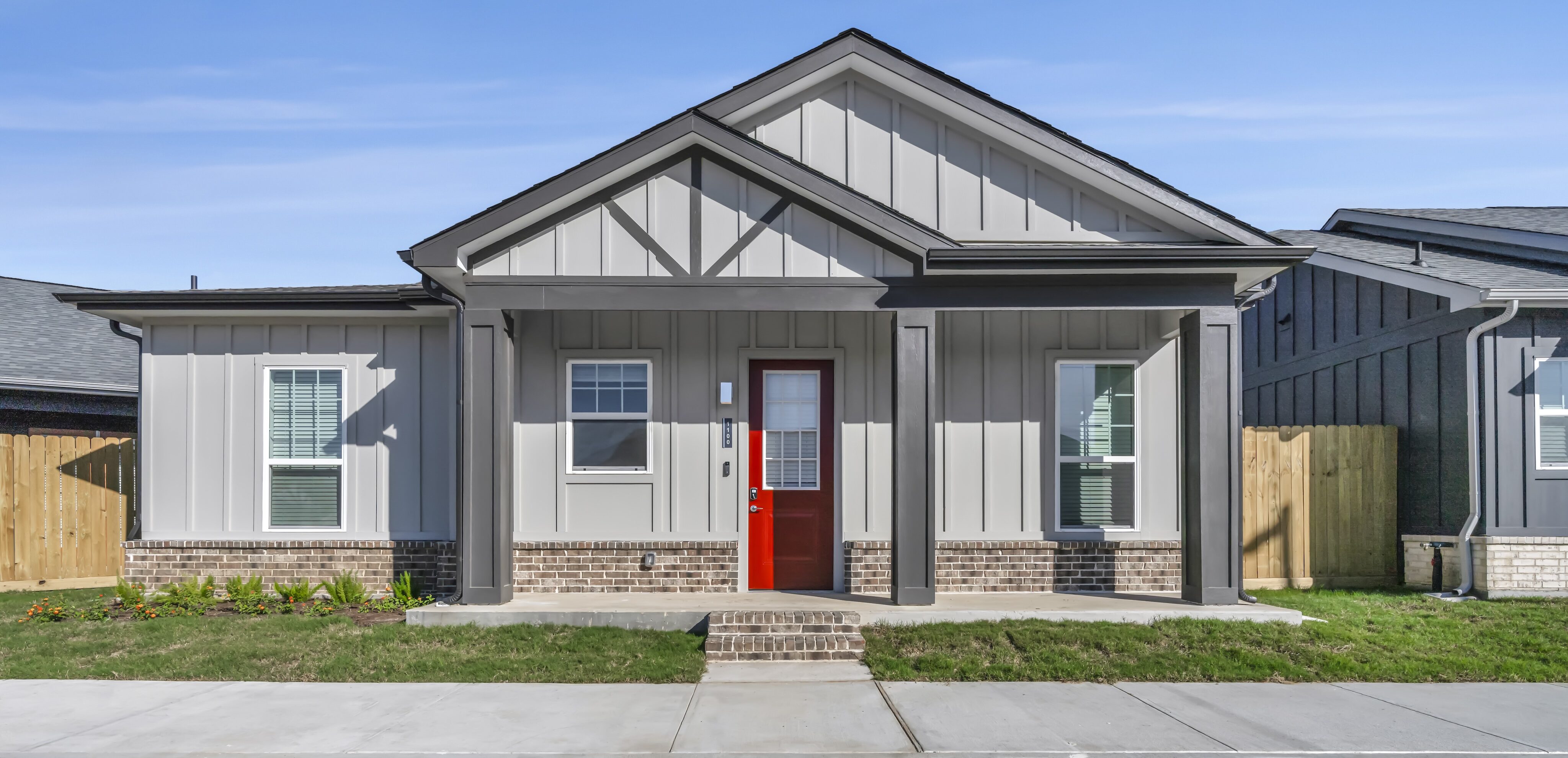 Single-story modern house with gray siding, brick accents, a red front door, white-trimmed windows, and a small front lawn, seen from a parking lot.