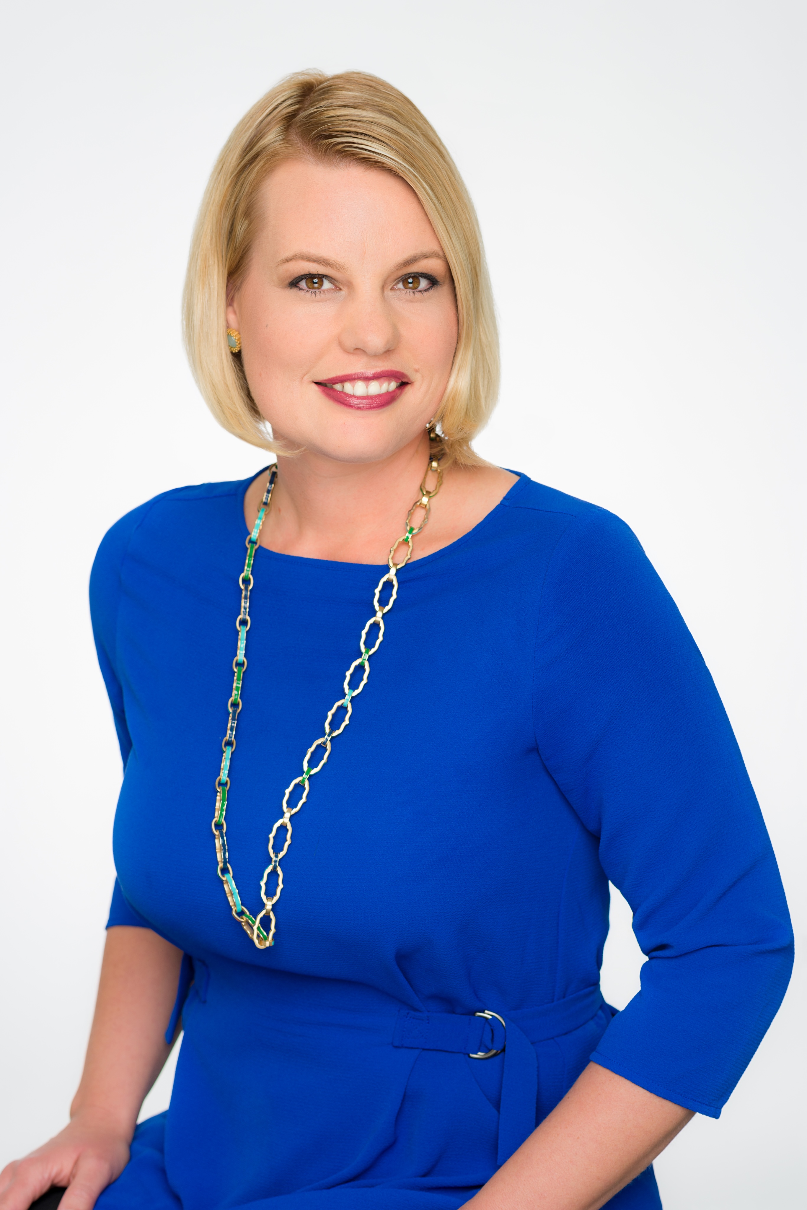 A woman with short blonde hair wearing a bright blue dress and a long chain necklace, smiling at the camera against a plain white background.
