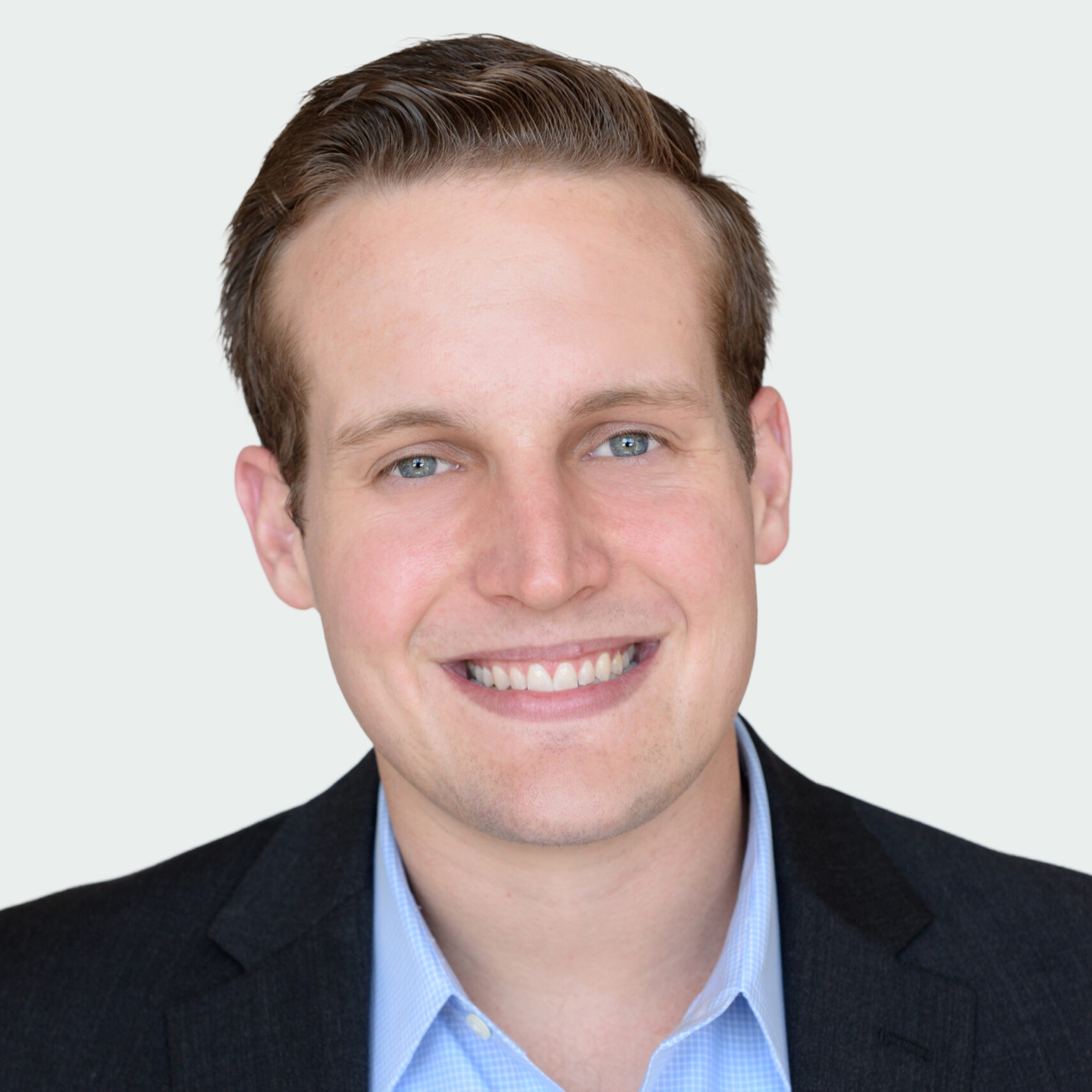 A man with short light brown hair, wearing a dark blazer over a light blue collared shirt, smiles at the camera against a plain light background.