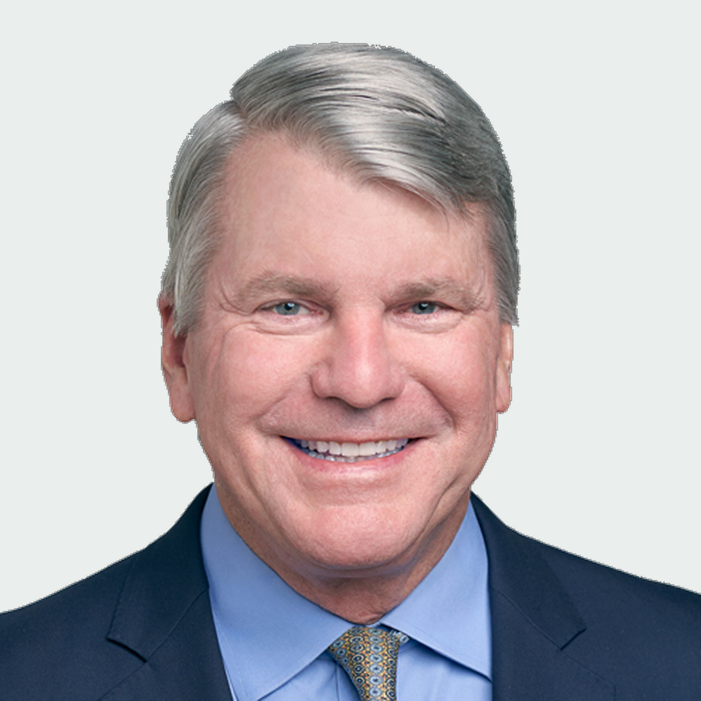 A middle-aged man with gray hair smiles while wearing a dark suit, blue shirt, and patterned tie against a plain background.