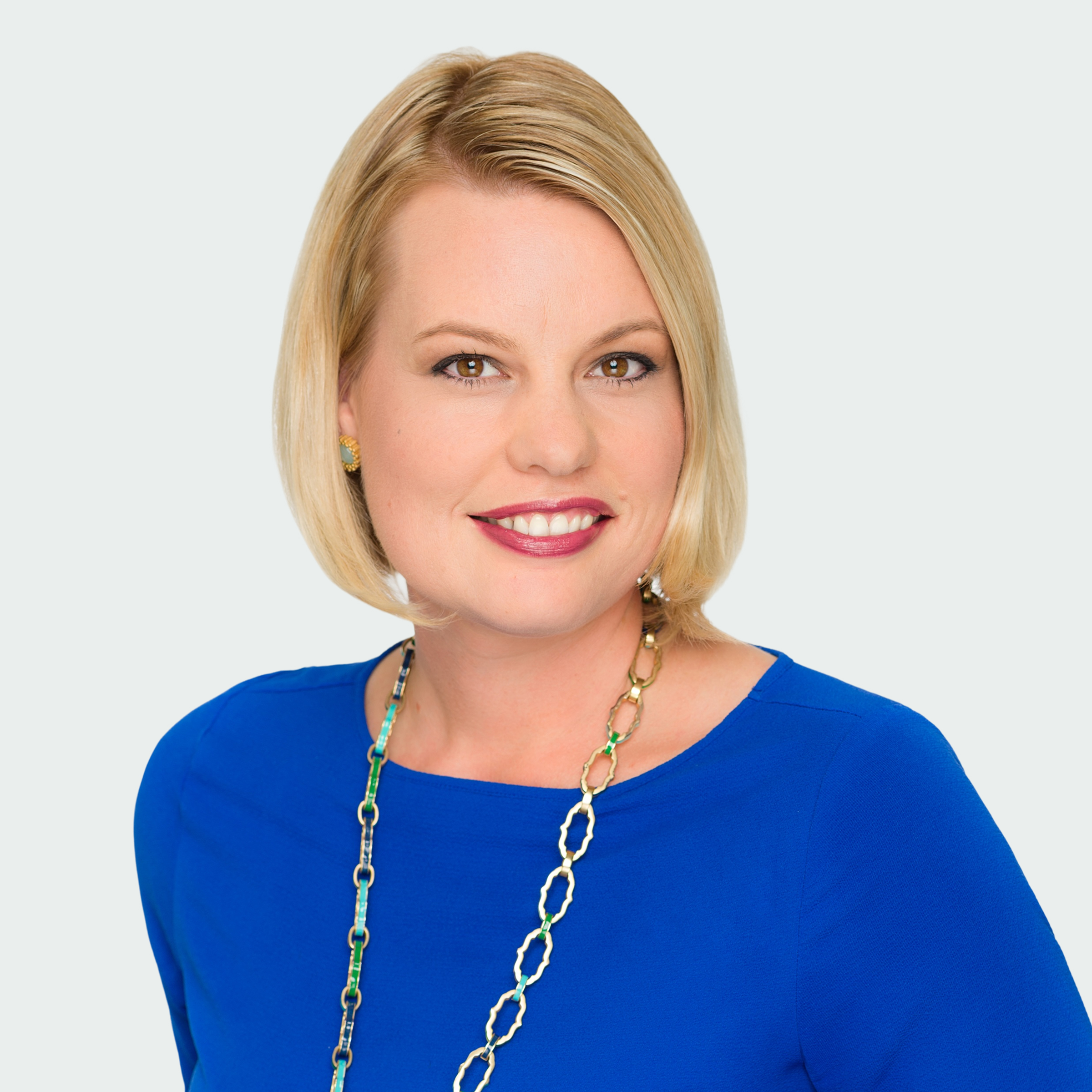 A woman with short blonde hair wearing a bright blue top and a long chain necklace, smiling at the camera against a plain light background.