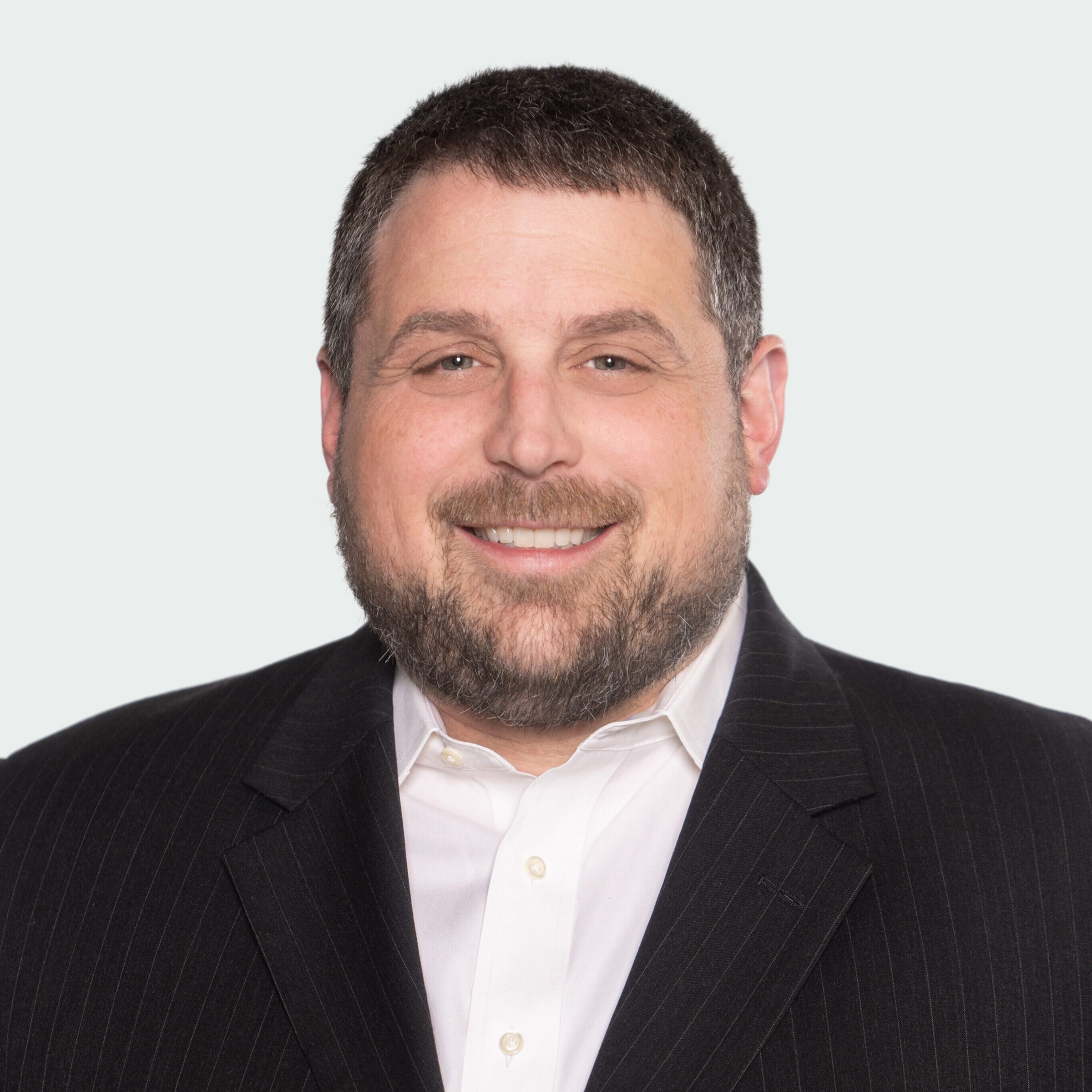 A man with short dark hair and a beard wears a black pinstripe suit jacket and white collared shirt, smiling at the camera against a plain light background.