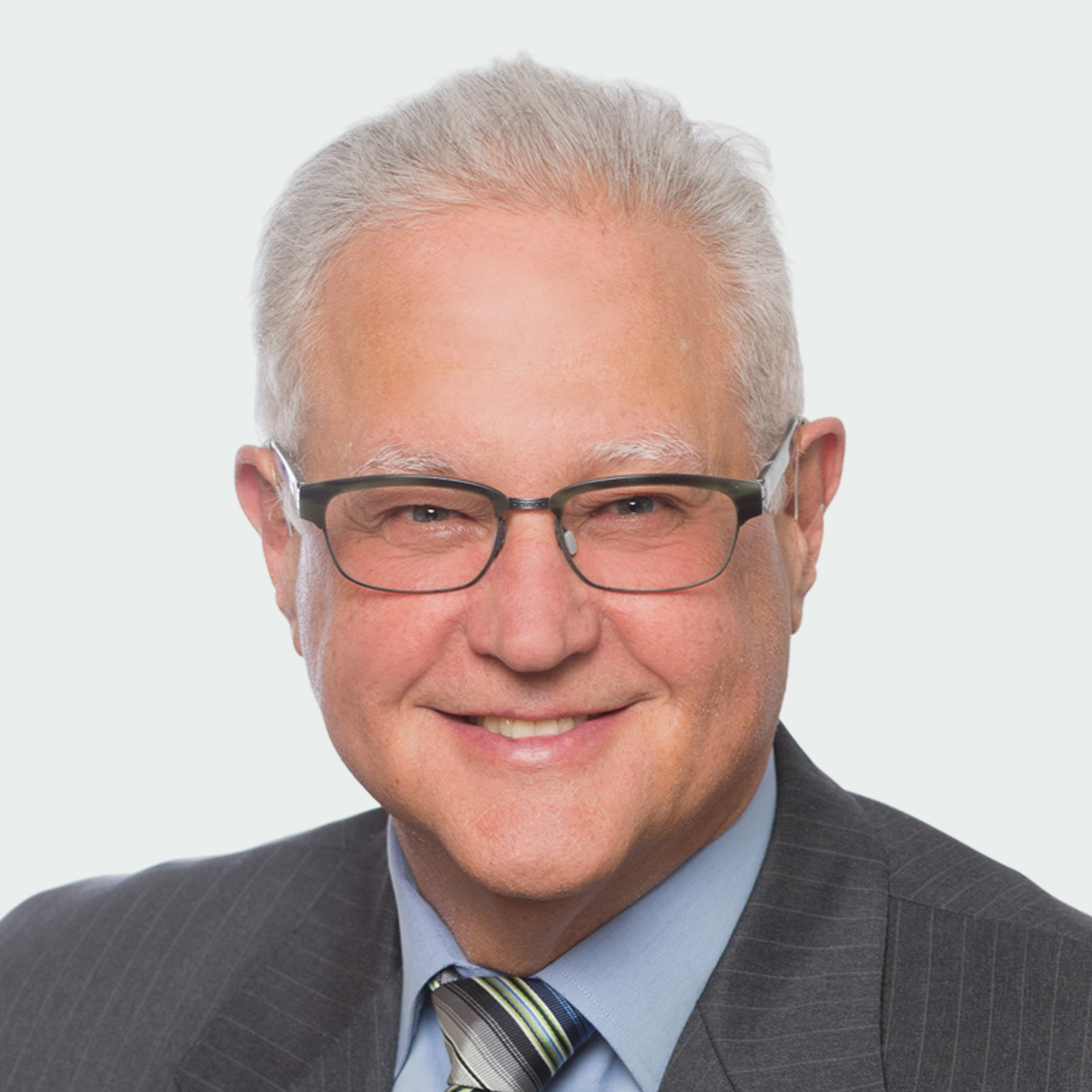 Smiling older man with gray hair and glasses wearing a striped suit, light blue shirt, and striped tie, posed against a plain white background.