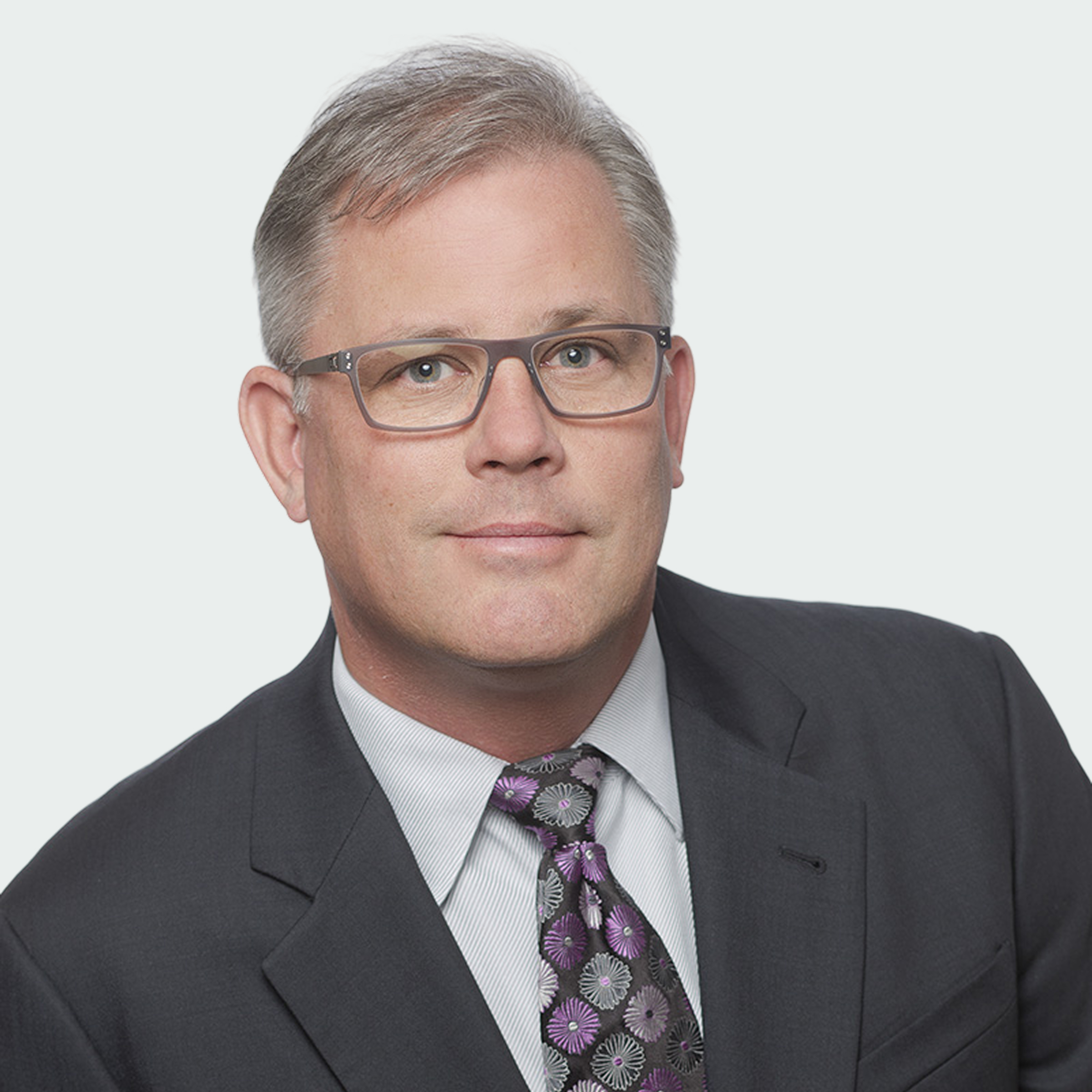 A man with gray hair and glasses wearing a dark suit, striped shirt, and patterned tie poses against a plain light background.
