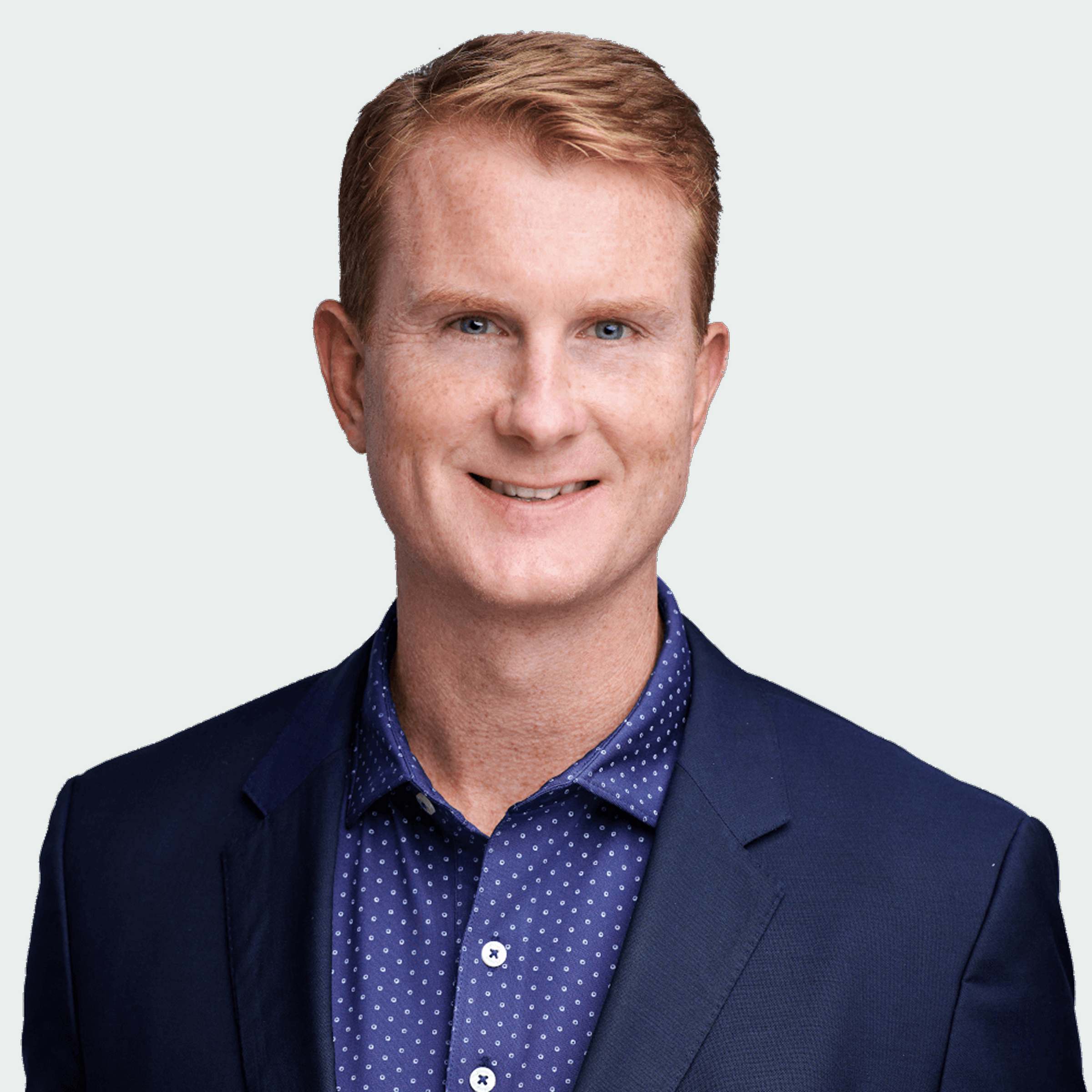 Man with short red hair wearing a blue suit jacket and a blue patterned shirt, smiling at the camera against a light gray background.