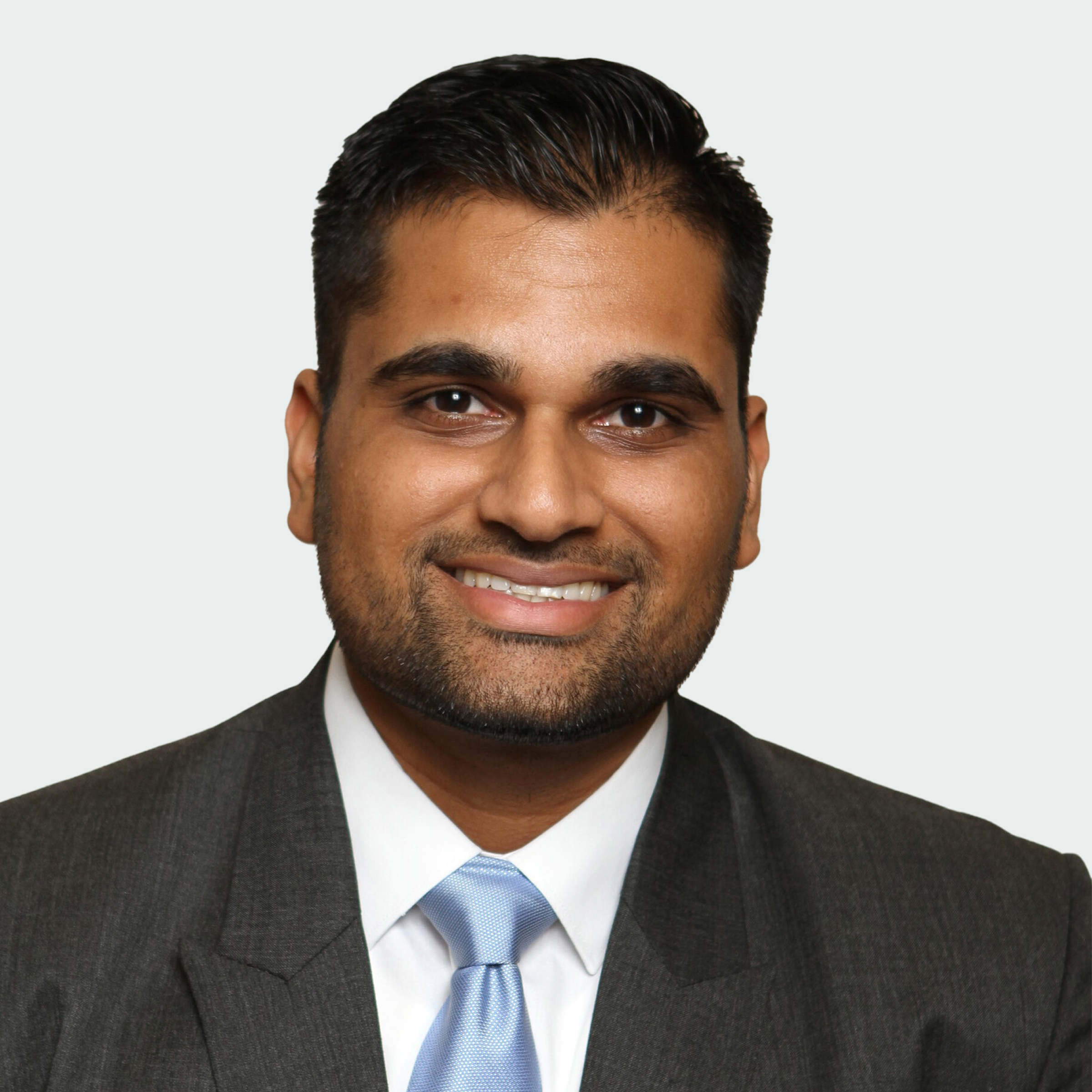 A man with short dark hair and a trimmed beard, wearing a gray suit, white shirt, and light blue tie, smiles at the camera against a plain light background.