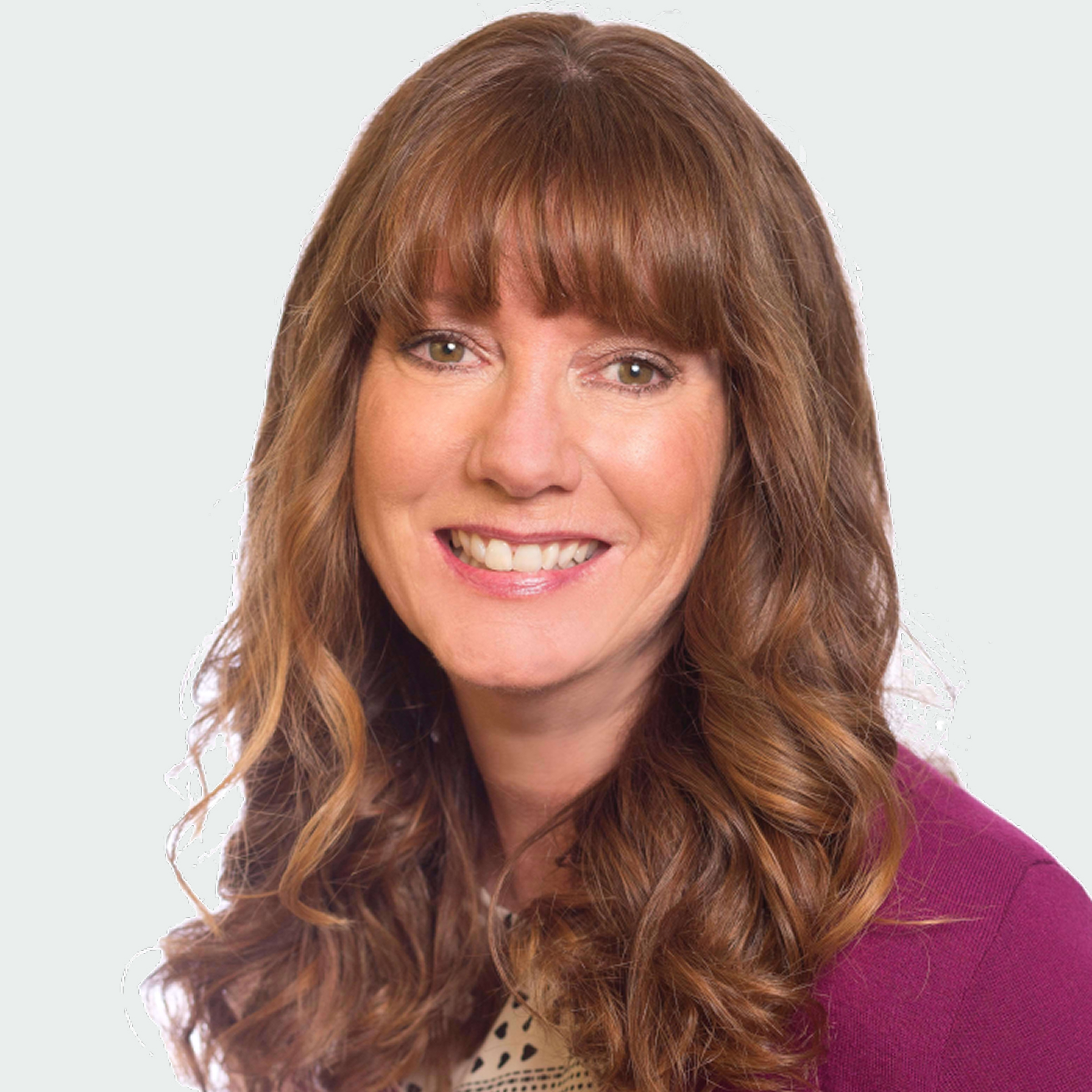 A woman with long, wavy brown hair and bangs is smiling at the camera. She is wearing a maroon jacket over a light-colored top against a plain background.
