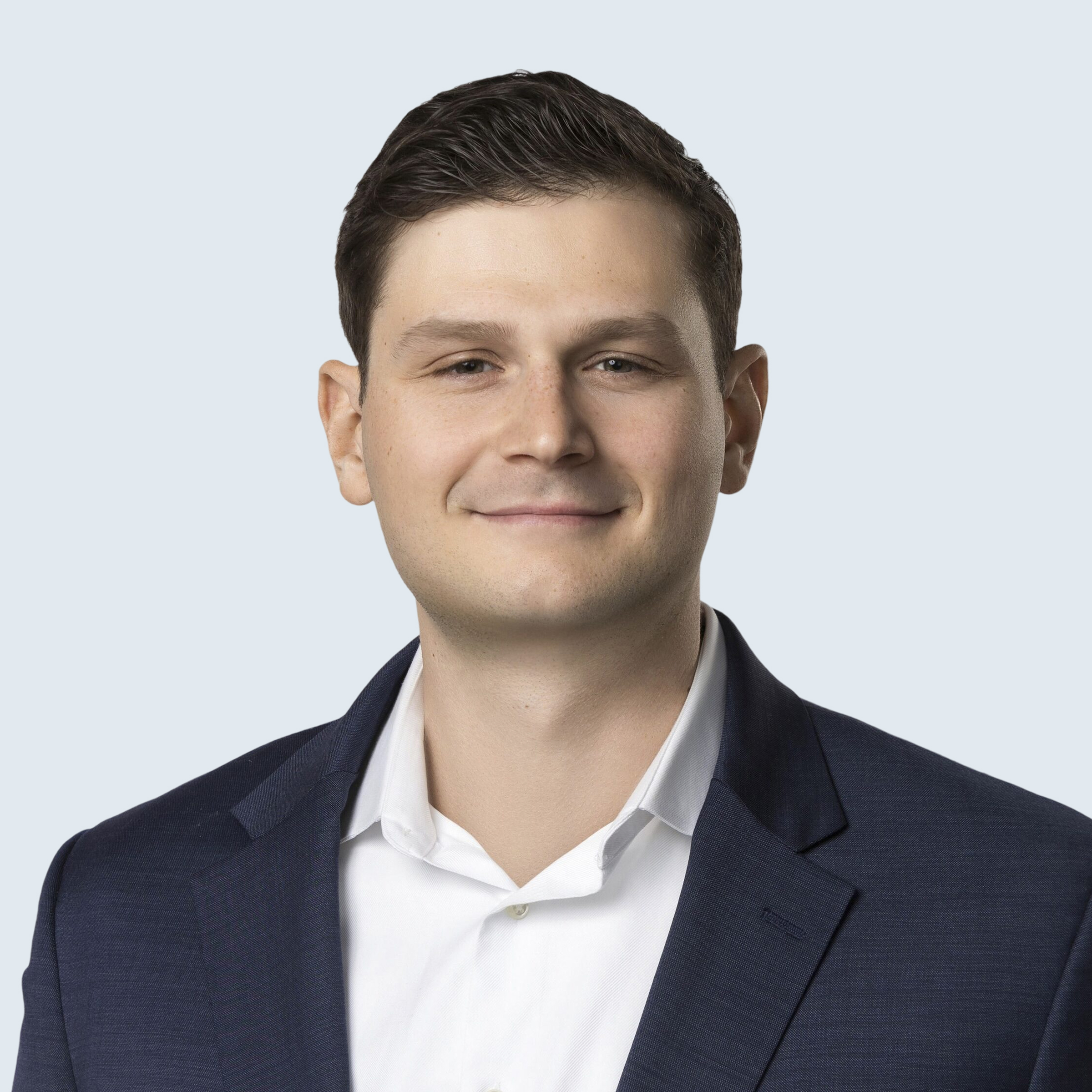 A man in a dark blue suit and white shirt smiles slightly at the camera against a light gray background.
