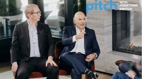 Two men in business attire sit and talk in front of a fireplace on a set labeled "elevator pitch with Jonathan & Mitch.